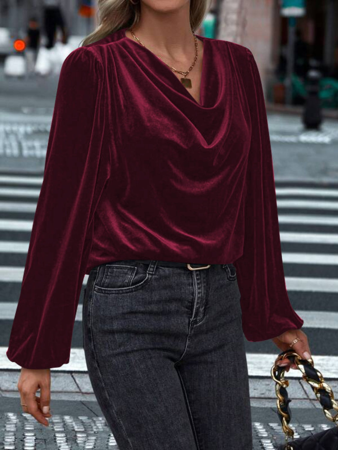 Model wearing a burgundy Lovelet cowl neck long sleeve blouse, paired with black jeans and a stylish chain handbag.